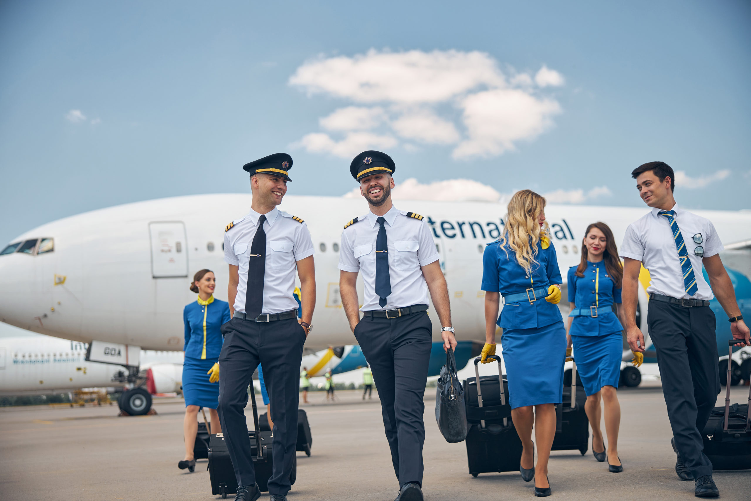 Des pilotes séduisants et de charmantes hôtesses de l'air discutent et sourient en trimballant des valises à l'aéroport avec un avion en arrière-plan.